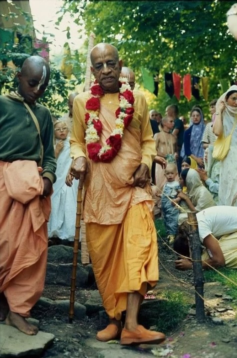 Srila Prabhupada walking in a garden in saffron robes with a red and white garland