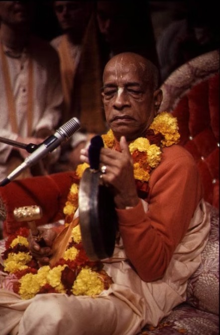 Srila Prabhupada seated at kirtan, striking a gong beside a microphone